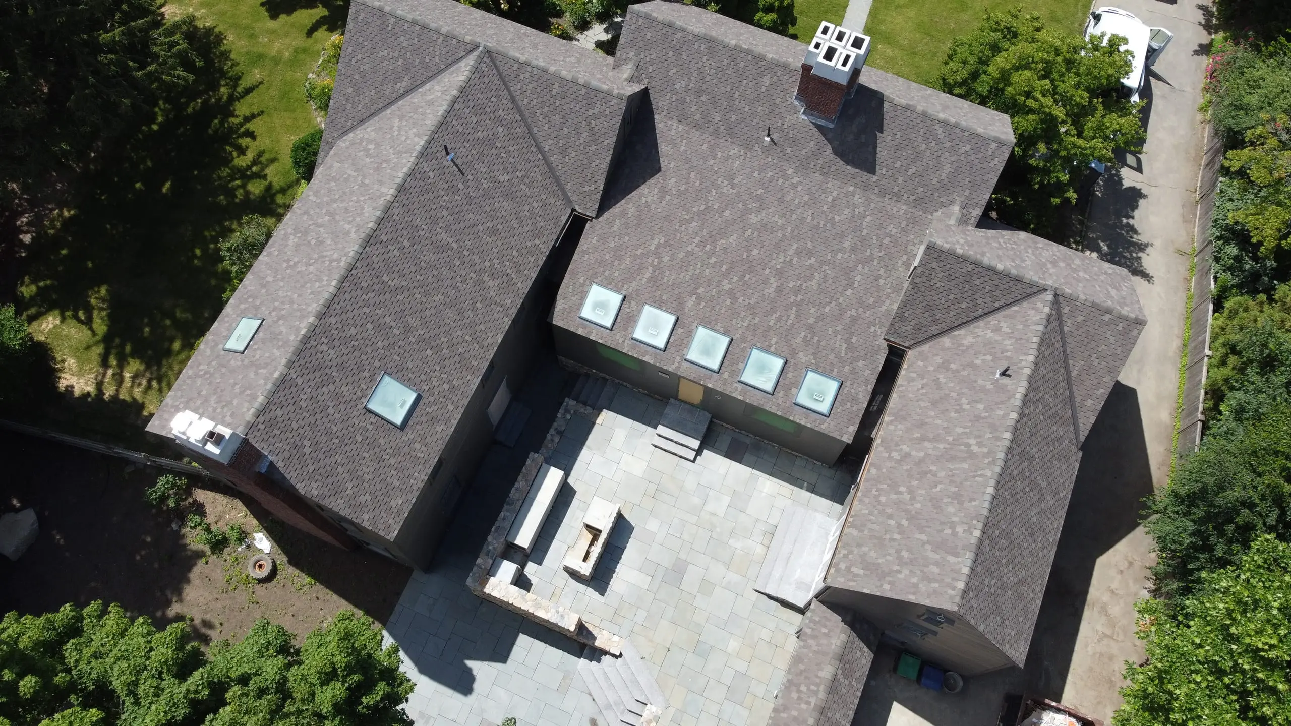 Aerial view of a completed asphalt shingle roof with five skylights and a brick chimney, over a bluestone courtyard
